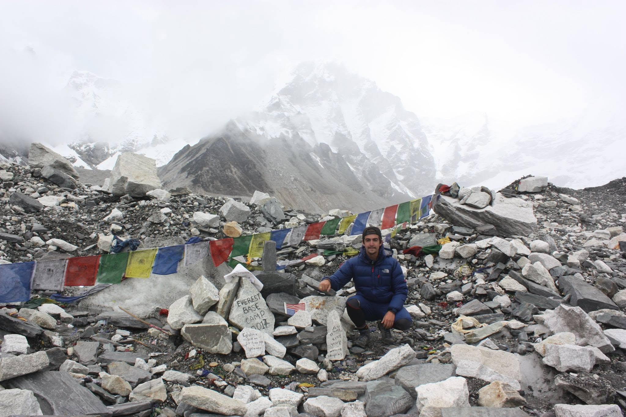 Alex at Everest Base Camp, surrounded by mountains and prayer flags